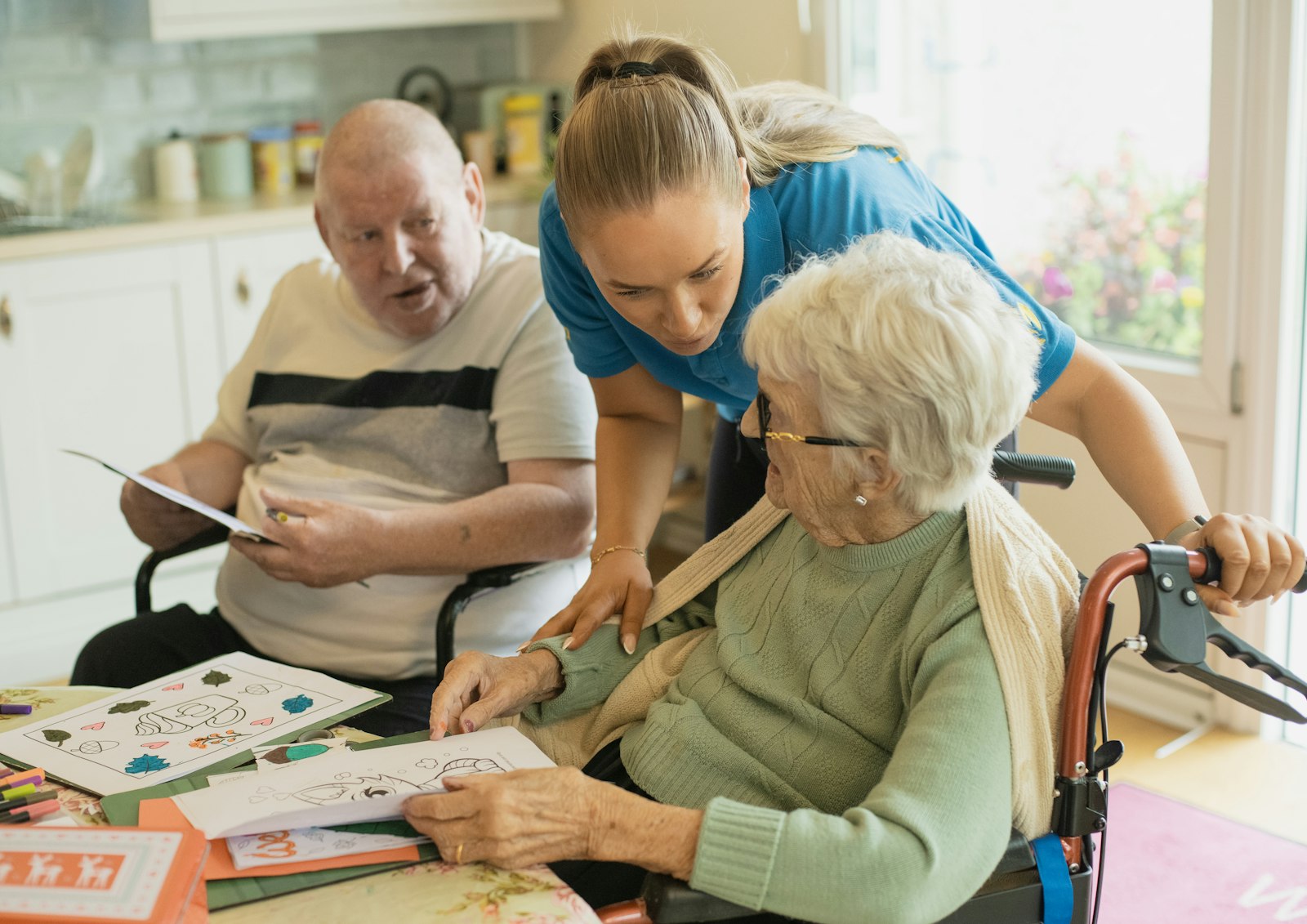 A caregiver leaning in to help an older woman with a drawing, beside an older man at the same table.
