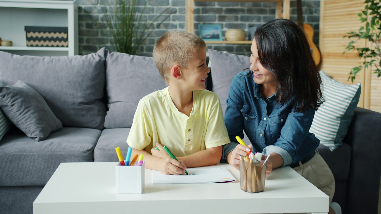 A mother and her young son drawing together at a coffee table.