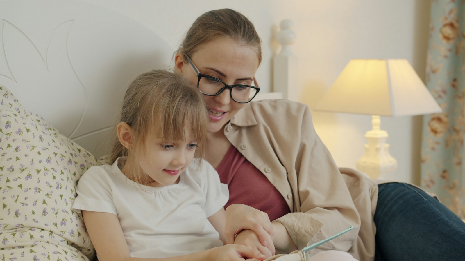 A woman with glasses reading a book with a young girl on a bed.