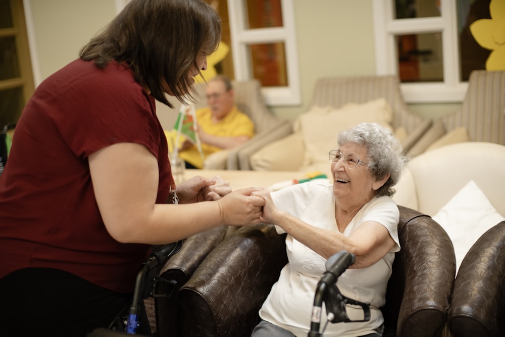 A caregiver holding an older woman's hands, helping her up from a chair.